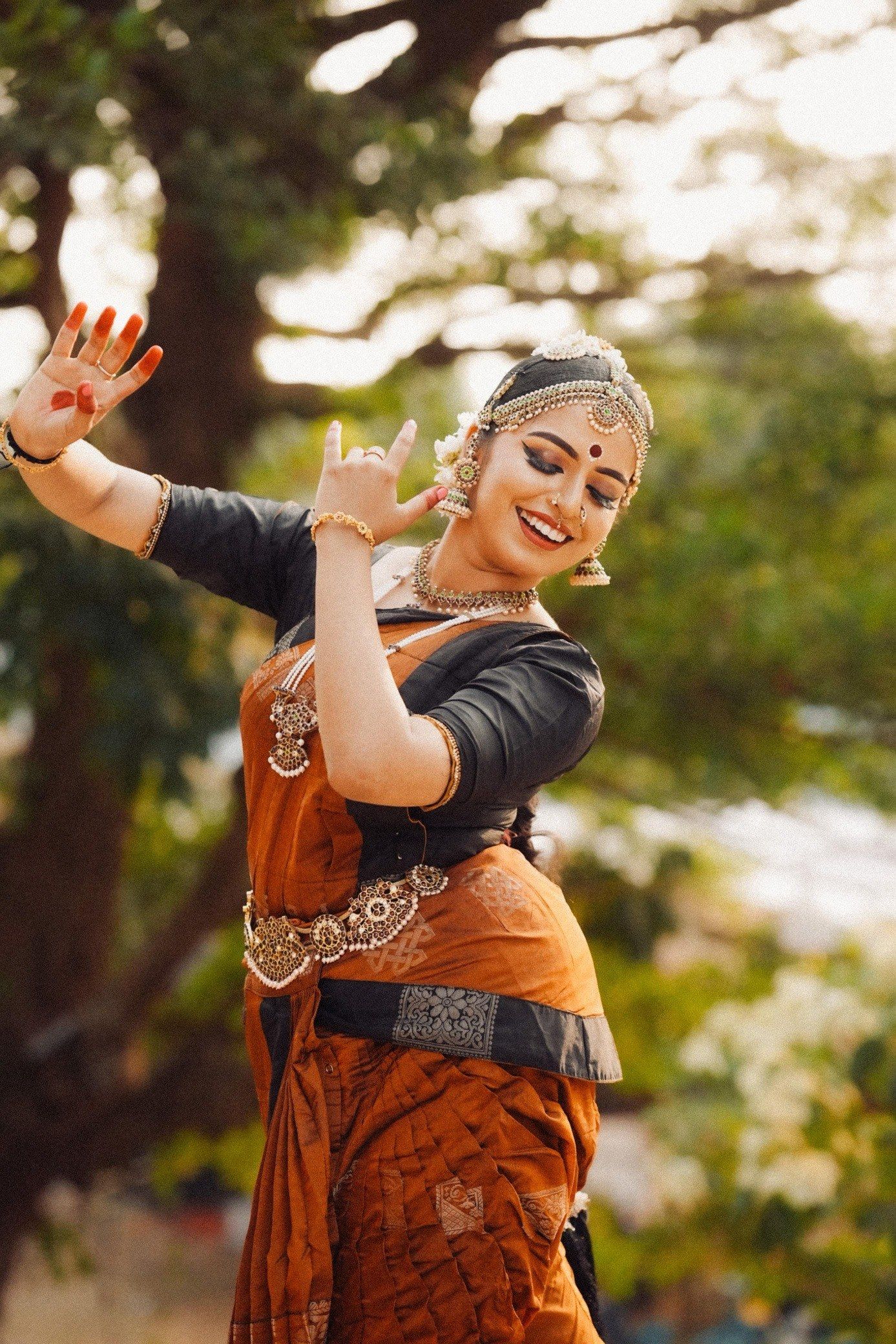 Medha Mayuri outdoors in an orange and blue costume, mid-gesture, trees behind her.