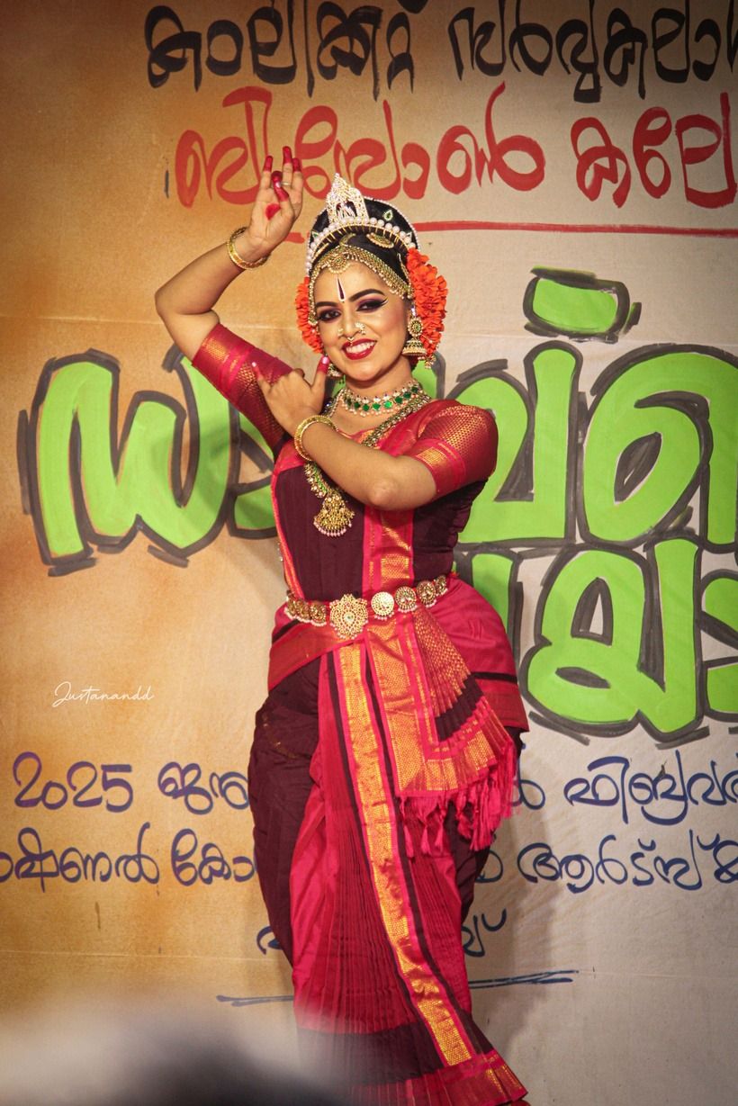 Medha Mayuri in a deep red costume performing in front of a competition banner.